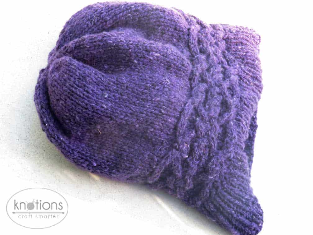 A purple worsted-weight knit hat laying flat. The hat has a ribbed brim, and a stockinette main body. Just above the ribbing, there is a celtic-knot style cable band that is knit horizontally. The top of the hat is gathered in slightly.